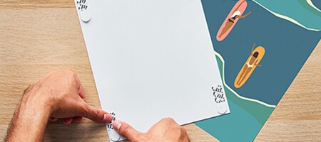 Un étudiant applique des Languettes adhésives pour posters Command™ au dos d’un poster afin d’accrocher celui-ci sur un mur de son logement étudiant
